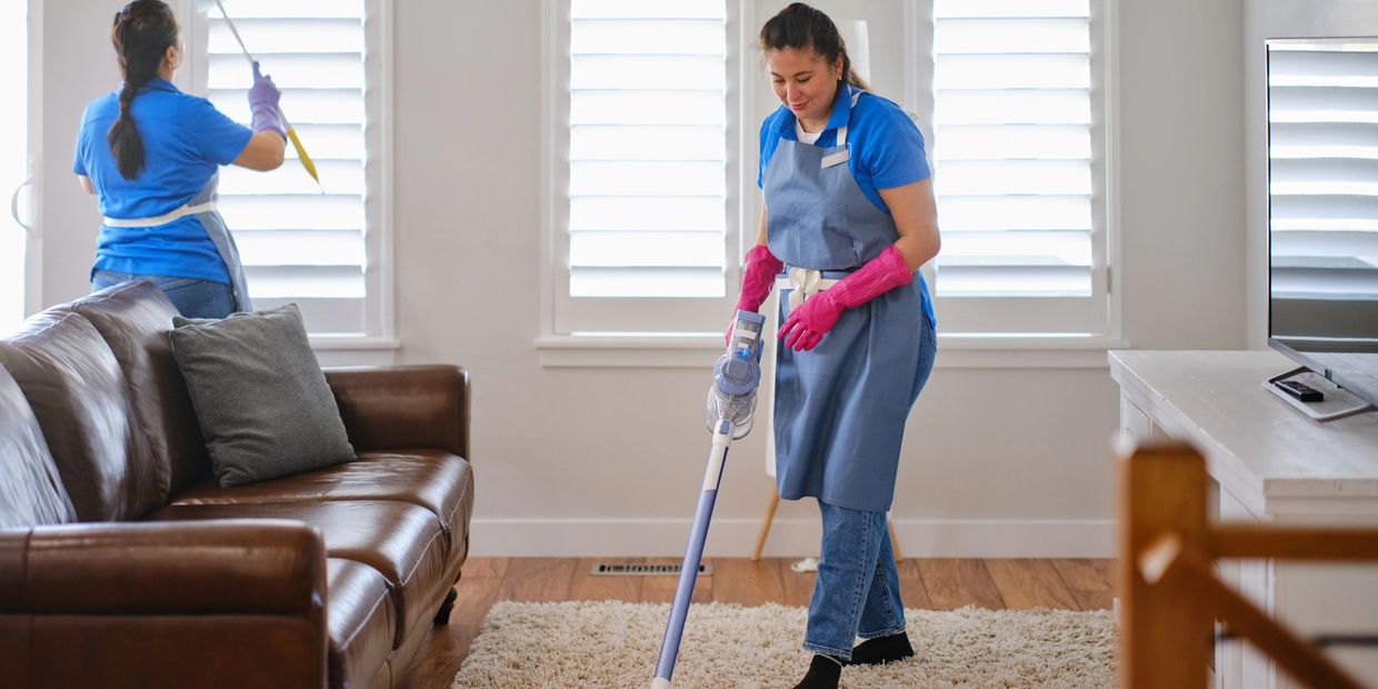 Two women cleaning a living room, vacuuming carpet and dusting windows.