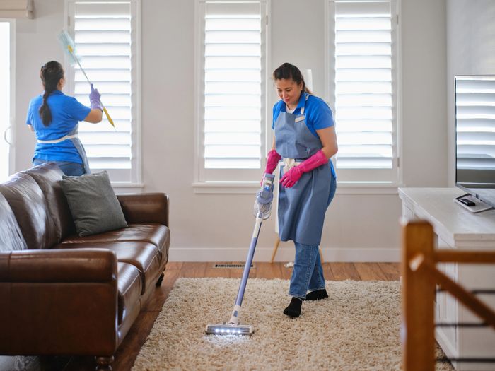 Two women cleaning a living room, vacuuming carpet and dusting windows.