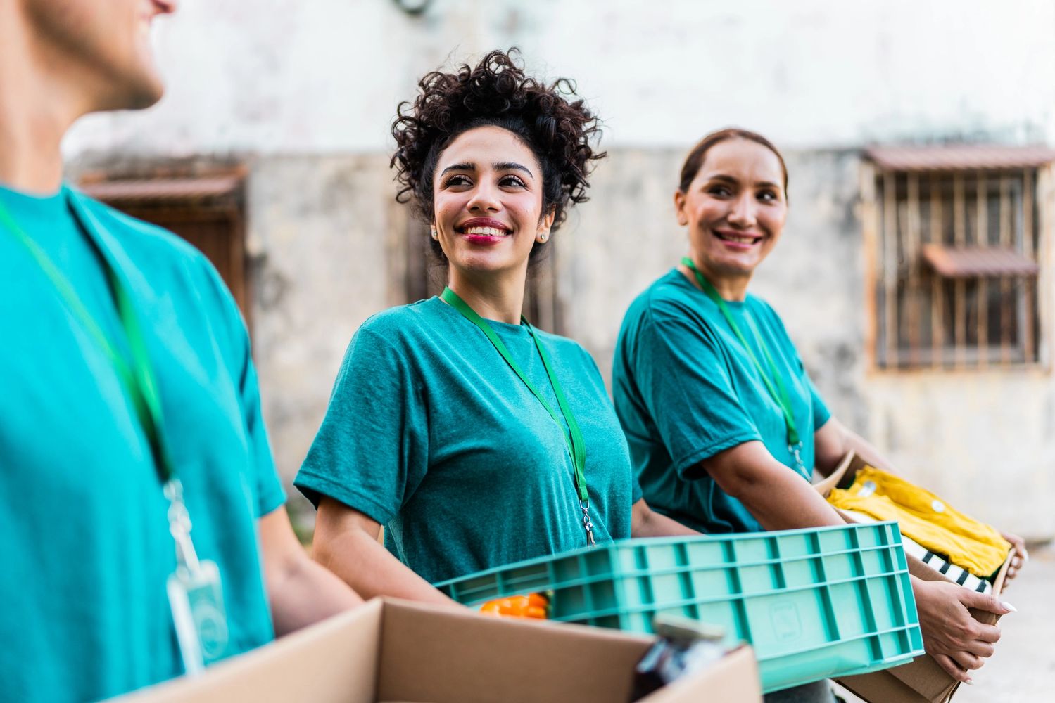 Volunteers in teal shirts smiling while holding boxes of donations outdoors.