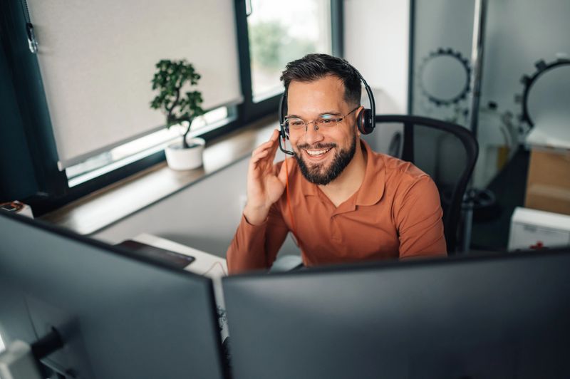 Customer service representative is working from his home office, smiling while using a headset and dual monitors to assist customers or clients