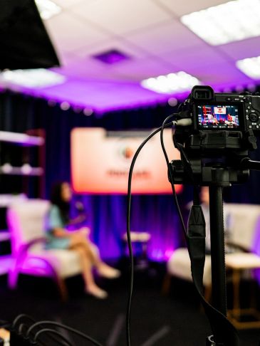 Camera filming a talk show set with purple lighting.