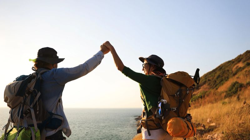 A Couple of adventure trekking hikers with backpacks walking uphill at sunset and arms raised for outdoor adventures.Mountain travel concept