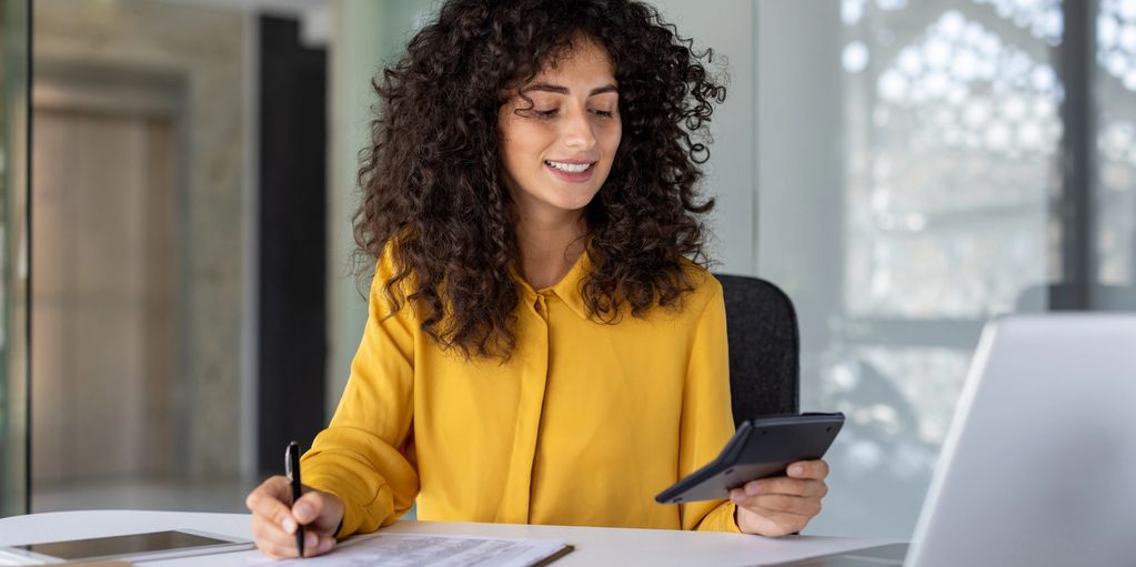 Woman in yellow shirt using a calculator and writing notes at her desk.