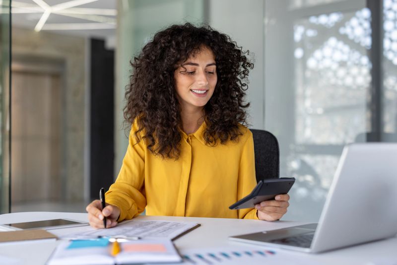 Latin American businesswoman accountant at office desk with calculator, pen, and laptop, demonstrating multitasking skills in financial work. Professional woman exemplifies confidence and efficiency.