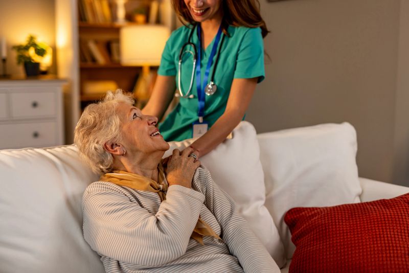 A nurse in scrubs and stethoscope provides compassionate support to an elderly woman sitting on a couch, symbolizing healthcare assistance and social connection in a comfortable home environment.