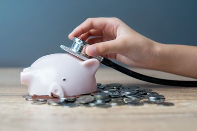 Hand holding stethoscope on piggy bank with coins, symbolizing financial health.