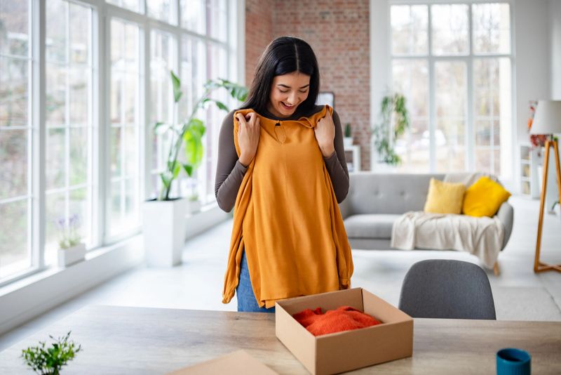 A woman lovingly examines a new orange blouse from her package, embodying happiness and surprise in a sunlit, modern home with a trendy interior style.