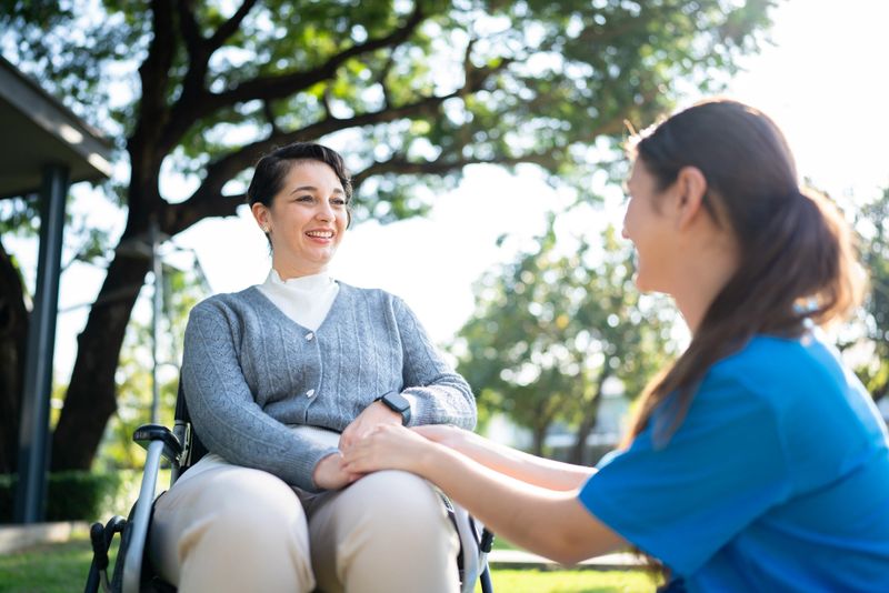 A nurse in blue scrubs holding hands with a smiling woman in a wheelchair, sharing a moment of care and connection in a sunny park.