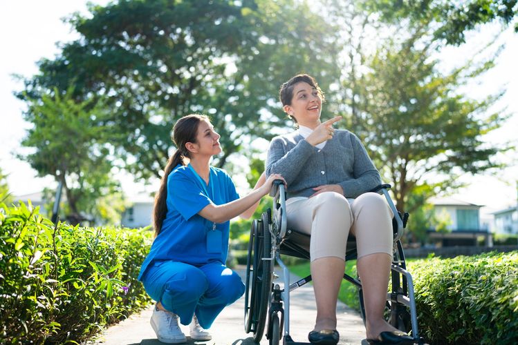 A nurse in blue scrubs assists a woman in a wheelchair outdoors.