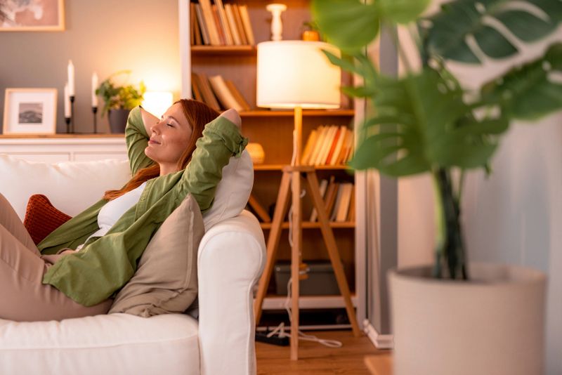 Redhead woman lying on a comfortable white sofa with hands behind her head, eyes closed, enjoying a serene moment of relaxation in a cozy, light-filled living room