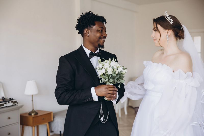 Elegant groom in tuxedo holding bouquet smiling at beautiful bride in white dress during interracial wedding ceremony
