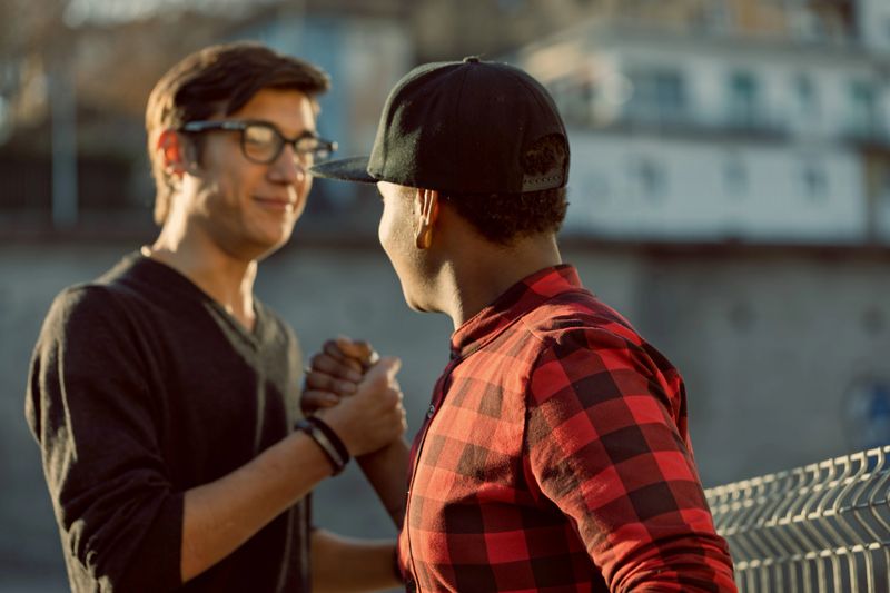 Two young men shaking hands in an urban setting, radiating friendship and respect while enjoying warm sunlight. Their casual attire and cheerful expressions highlight a genuine connection