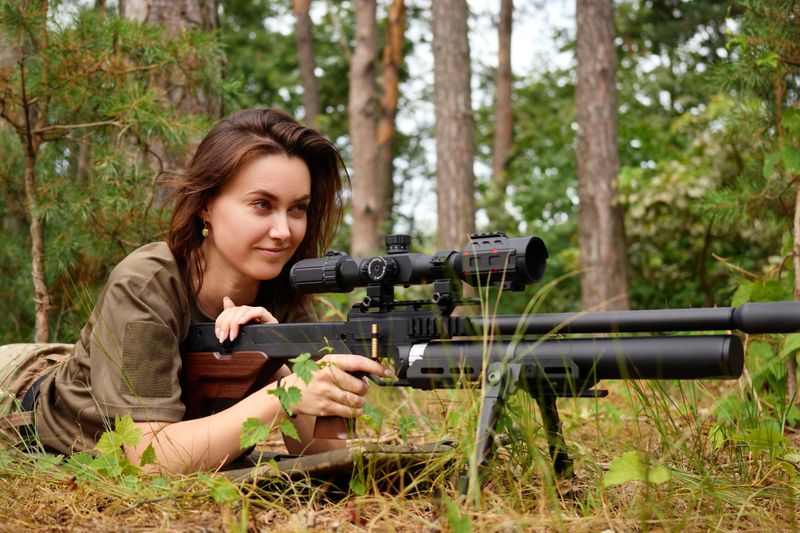 A focused woman lies on the ground, aiming a sniper rifle equipped with a scope in a wooded area. Tall trees surround her as she practices her shooting skills.