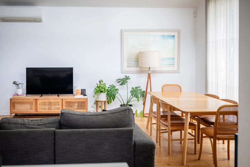 A sophisticated open-plan layout combining a living room and dining area with ample lighting, featuring a grey couch, wooden furniture, and a lush green plant decor.