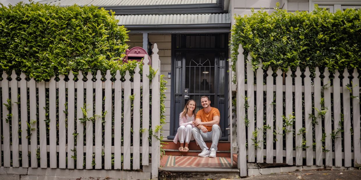 A happy couple sitting on the porch steps of their charming home with a white picket fence.