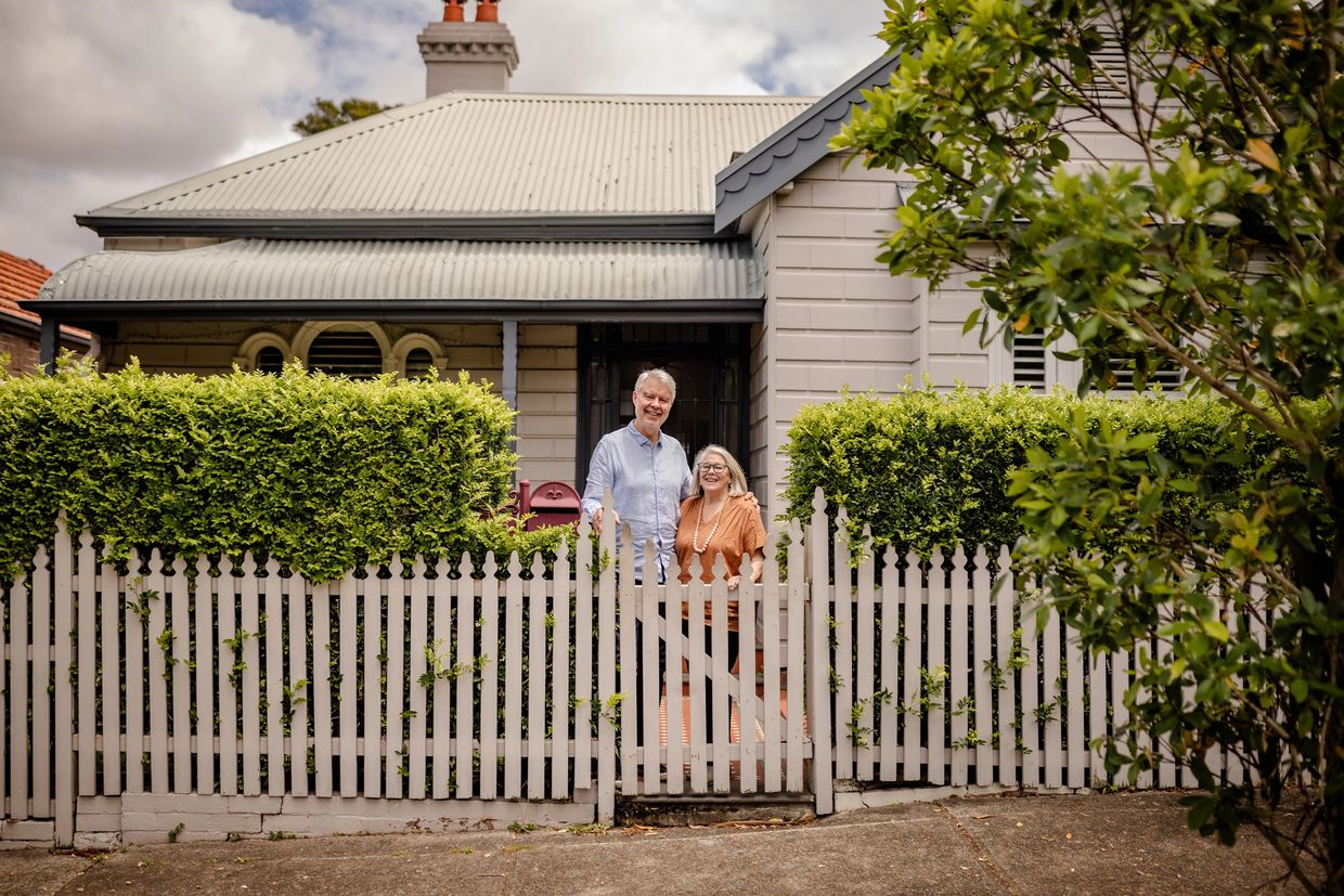 Happy elderly couple standing behind a white picket fence in front of their house.