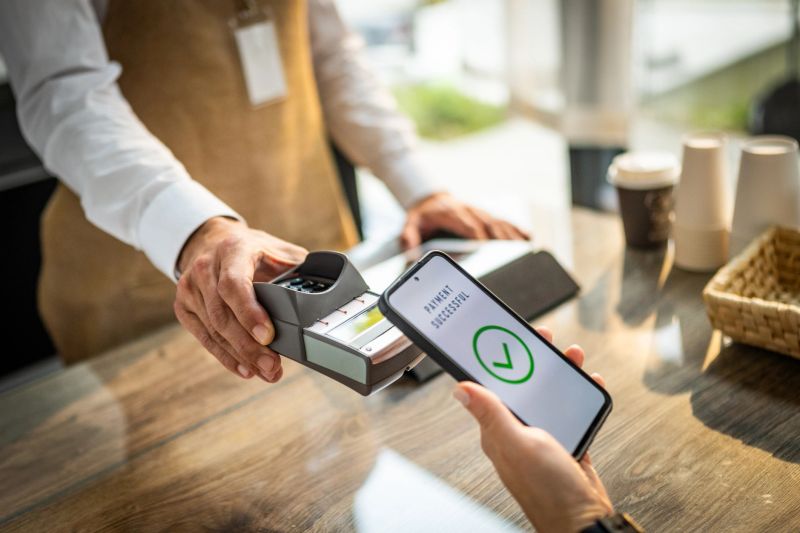 An unrecognizable male customer uses a smartphone to make a contactless payment to a barista at a coffee shop, showcasing modern technology and convenient transactions.
