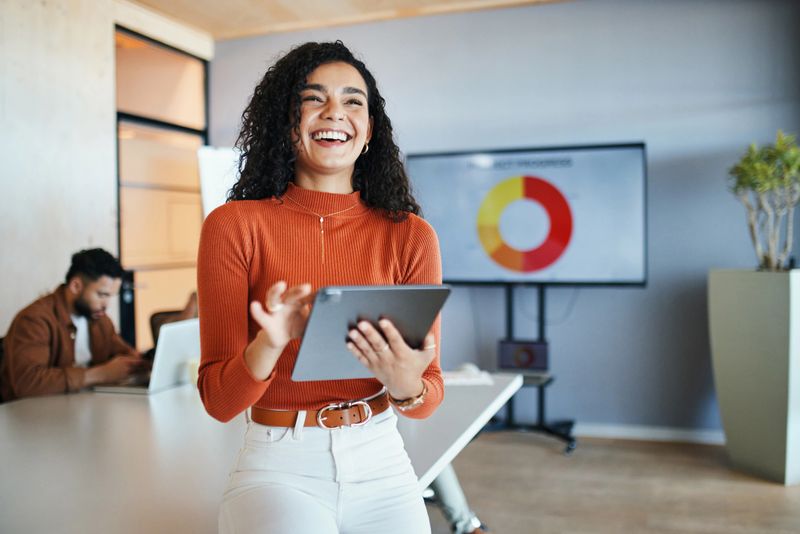 Smiling businesswoman with curly hair presents data on a tablet. A pie chart is displayed on a screen behind her. A colleague is working in the background, creating a collaborative office environment.