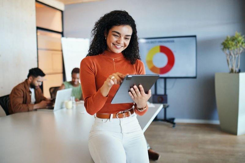 A smiling woman in an orange turtleneck and white pants interacts with a tablet in a modern office. Blurred colleagues work in the background, with a presentation screen showing a pie chart visible.