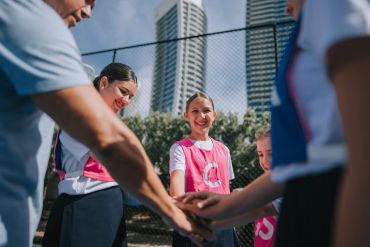 Young girls in sports jerseys join hands in a team huddle outdoors.