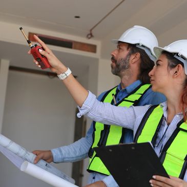 Two construction workers inspecting a ceiling with a walkie-talkie and blueprints.