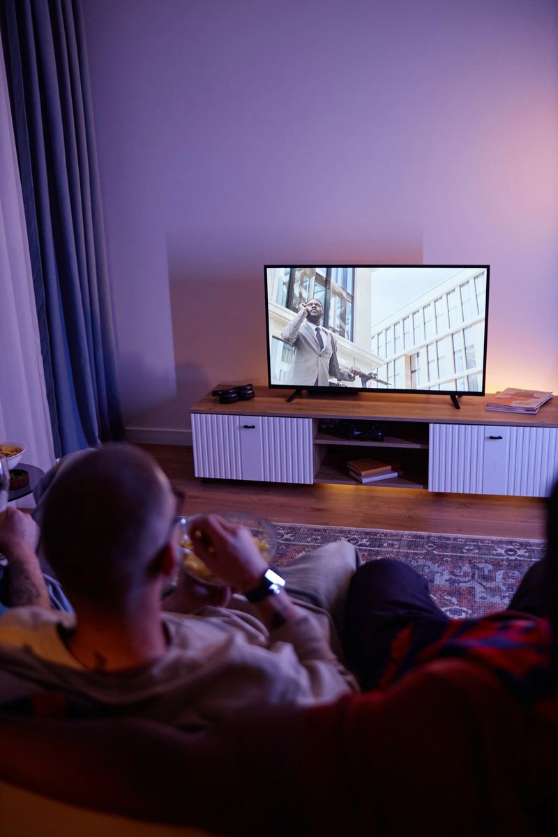 Vertical back view of friends watching Tv at night in dark room while sitting on sofa with focus on TV screen, copy space