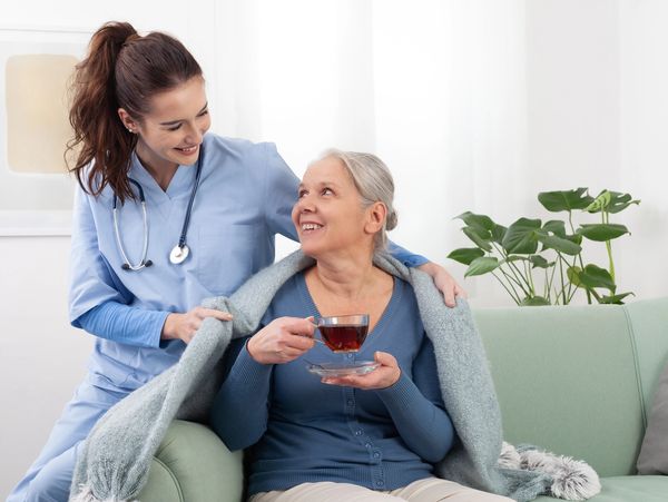 Female caregiver, wearing scrubs and a stethoscope, placing a blanket around a smiling elderly woman
