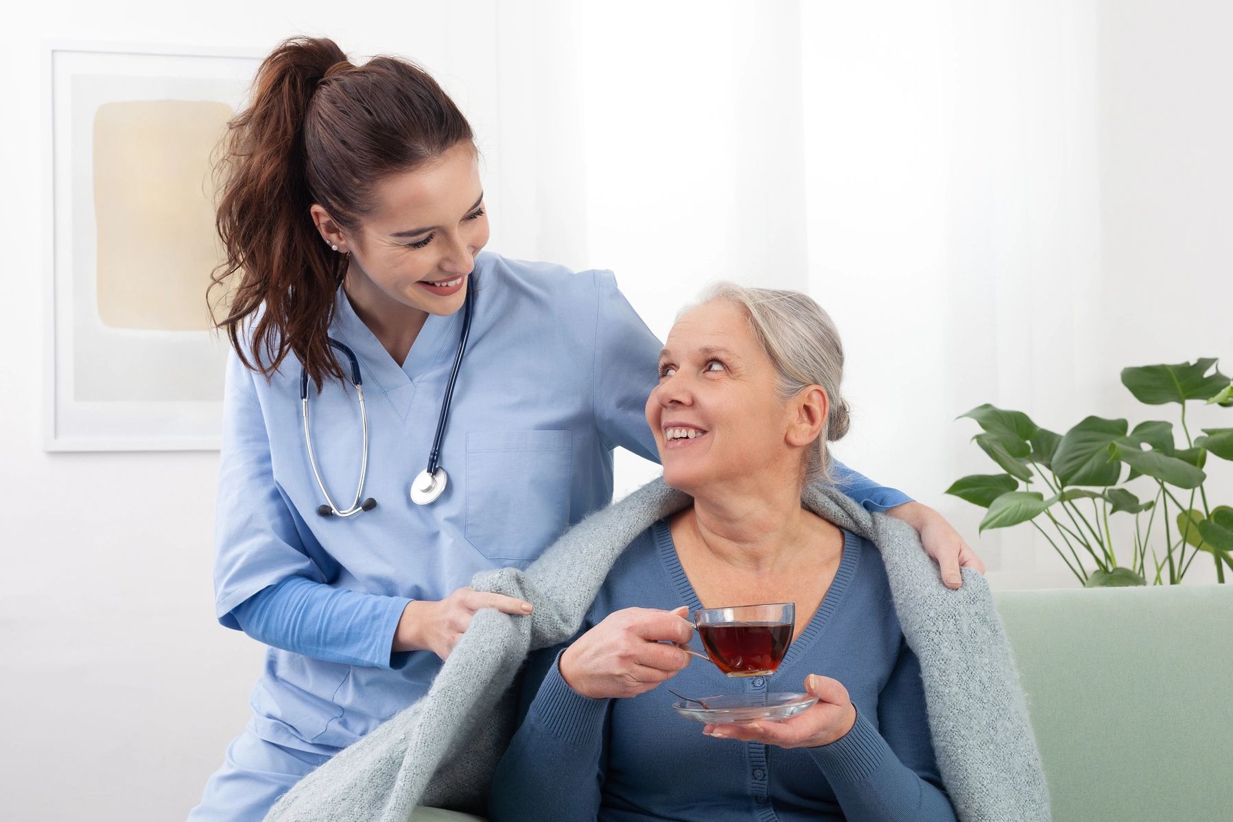 Nurse warmly covering elderly woman with a blanket while she enjoys tea.