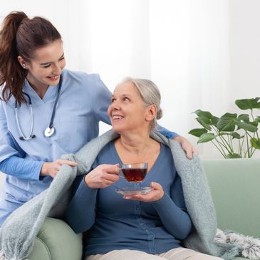 Nurse warmly covering elderly woman with a blanket while she enjoys tea.