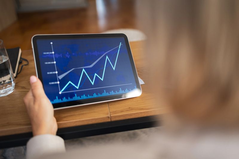 A close-up of a person holding a digital tablet displaying an upward-trending financial graph, symbolizing data analysis and business growth. The scene evokes a sense of modern technology and financial insight.