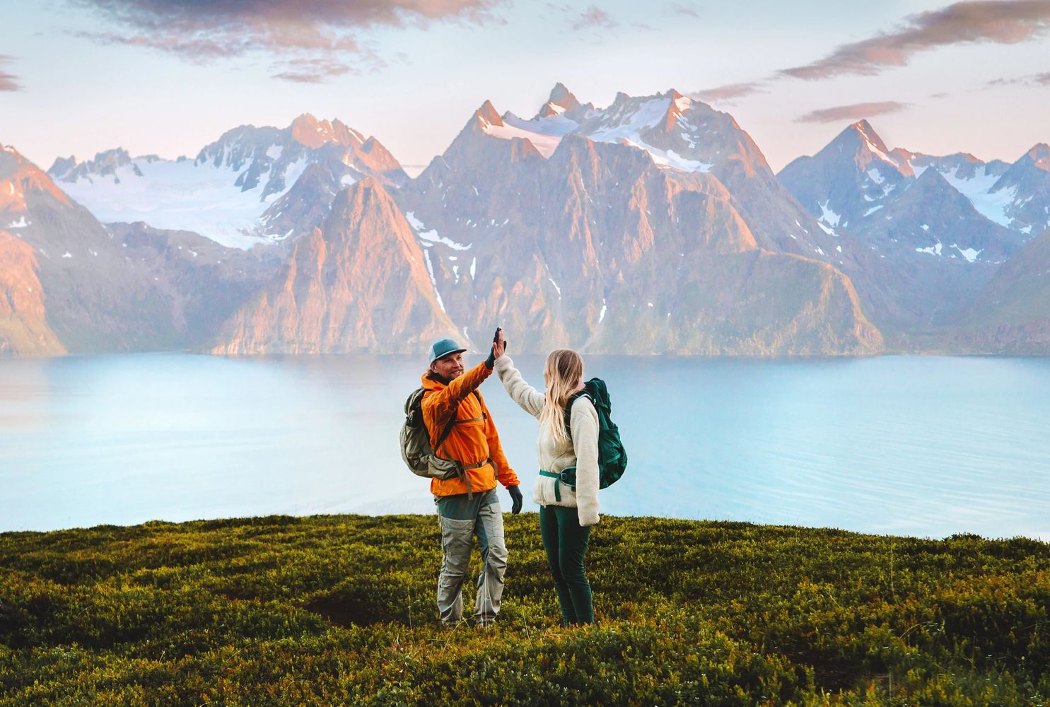 Two hikers high-five on a green hill with snowy mountains and a lake in the background.
