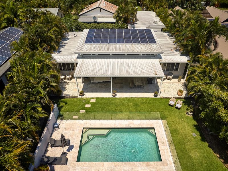 Unoccupied swimming pool surrounded by glass, manicured lawn, lush green foliage, covered patio with furniture, and solar panels on roof. New South Wales.
