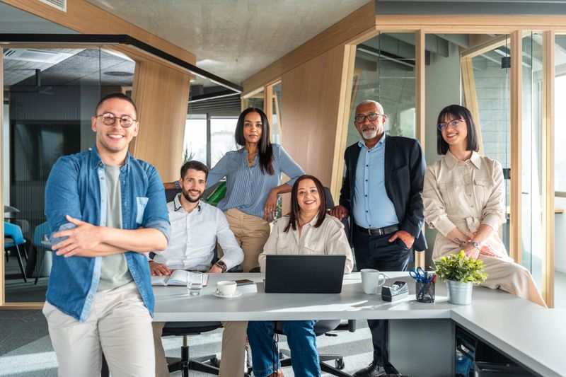 A confident group of professional men and women of varying ethnic backgrounds collaborate around a laptop in a modern workspace, dressed in business casual attire.
