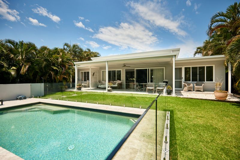 View across swimming pool surrounded by glass, manicured lawn, patio with furniture, and sliding door access to interior.