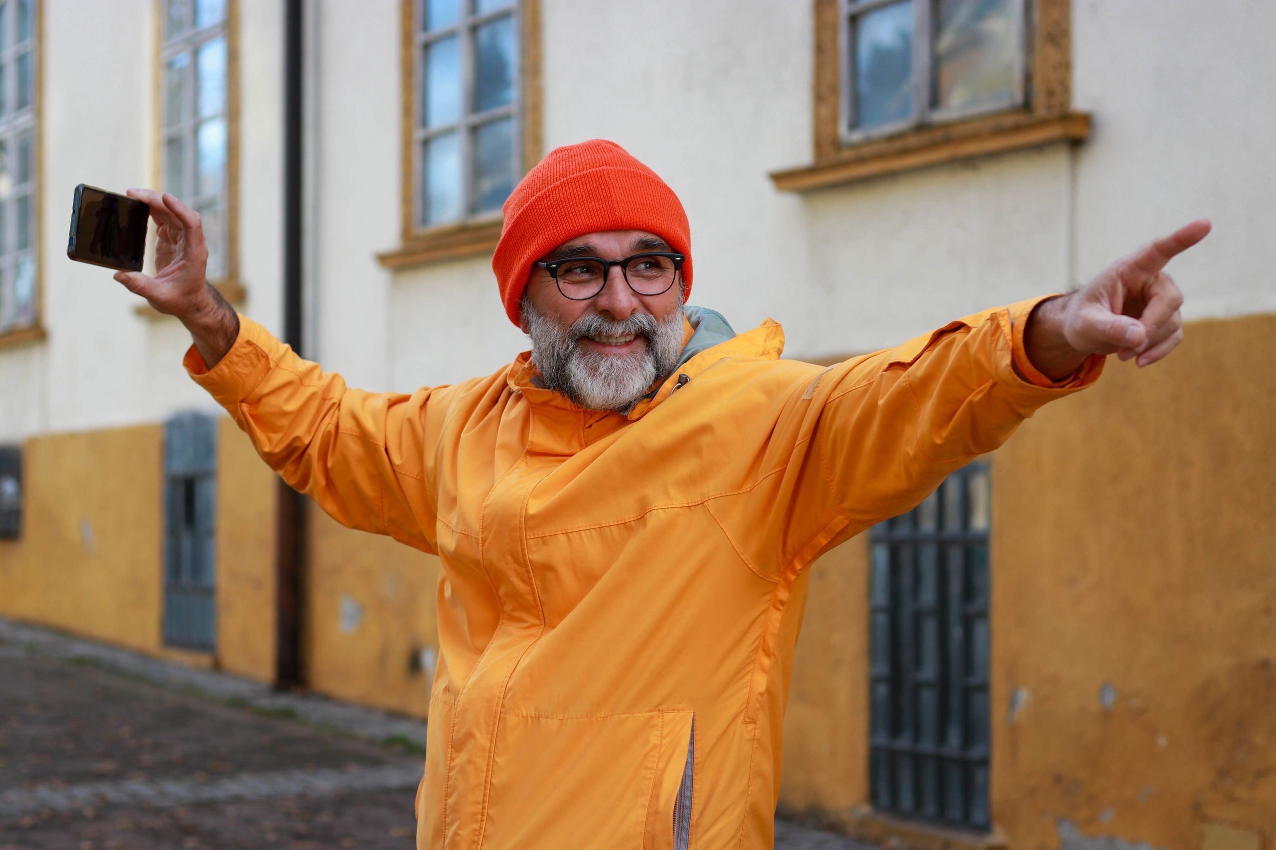 Smiling man in orange jacket and beanie holding phone, pointing excitedly outdoors.