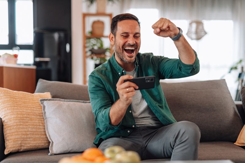 Happy man sitting comfortably on the sofa, playing games on his mobile phone at home, celebrating his victory with excitement and joy as he achieves success in the game