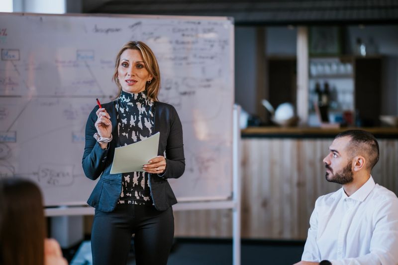 A professional woman presents in front of colleagues during a collaborative business meeting. She gestures while explaining ideas, creating an engaging and dynamic workplace atmosphere. A whiteboard with diagrams is in the background, adding depth and illustrating the context of the workplace. This captivating image highlights teamwork, professionalism, and leadership in the corporate environment.