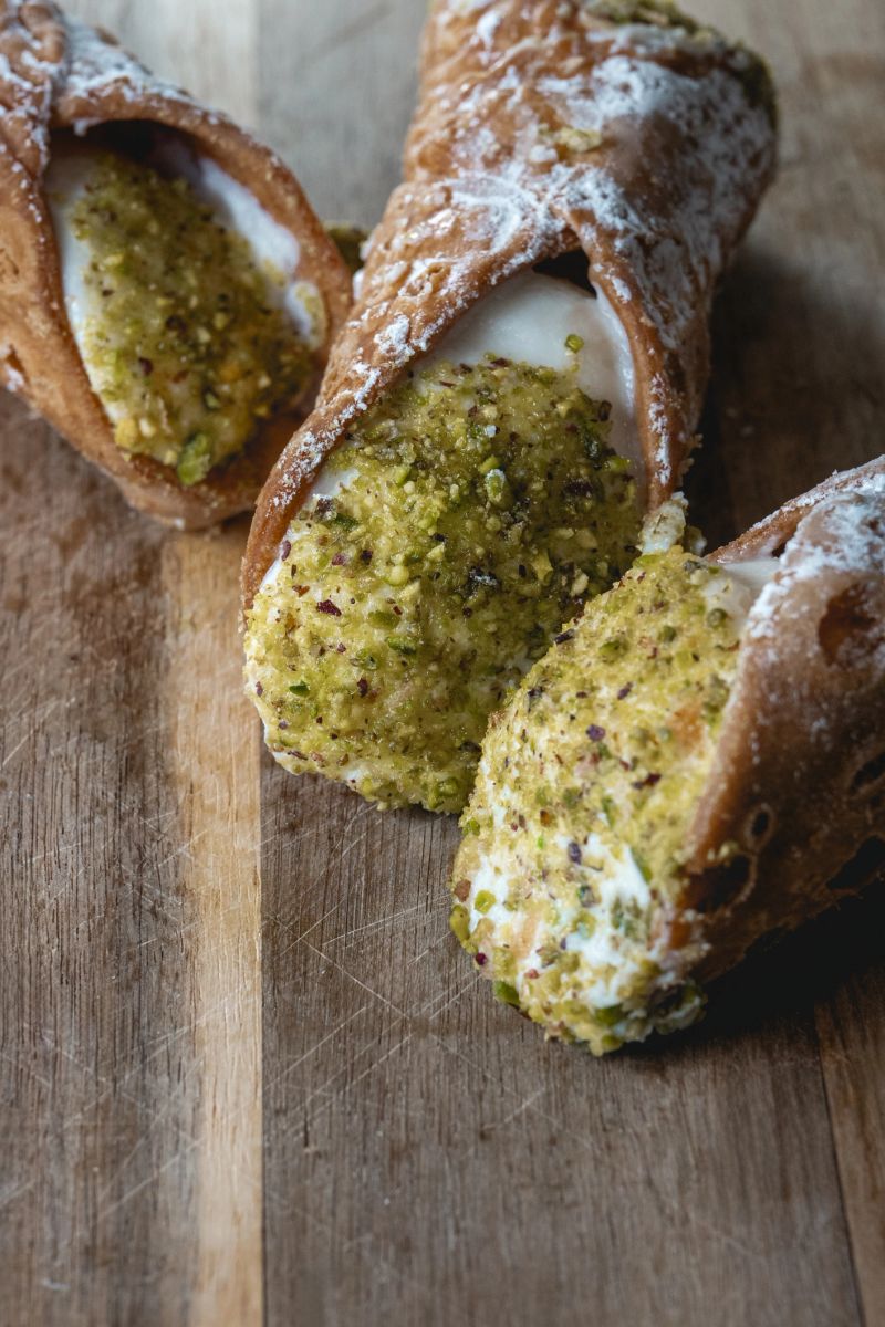 Close-up of traditional Sicilian cannoli filled with ricotta cream and topped with crushed pistachios, placed on a rustic wooden board. A perfect example of Italian dessert craftsmanship.