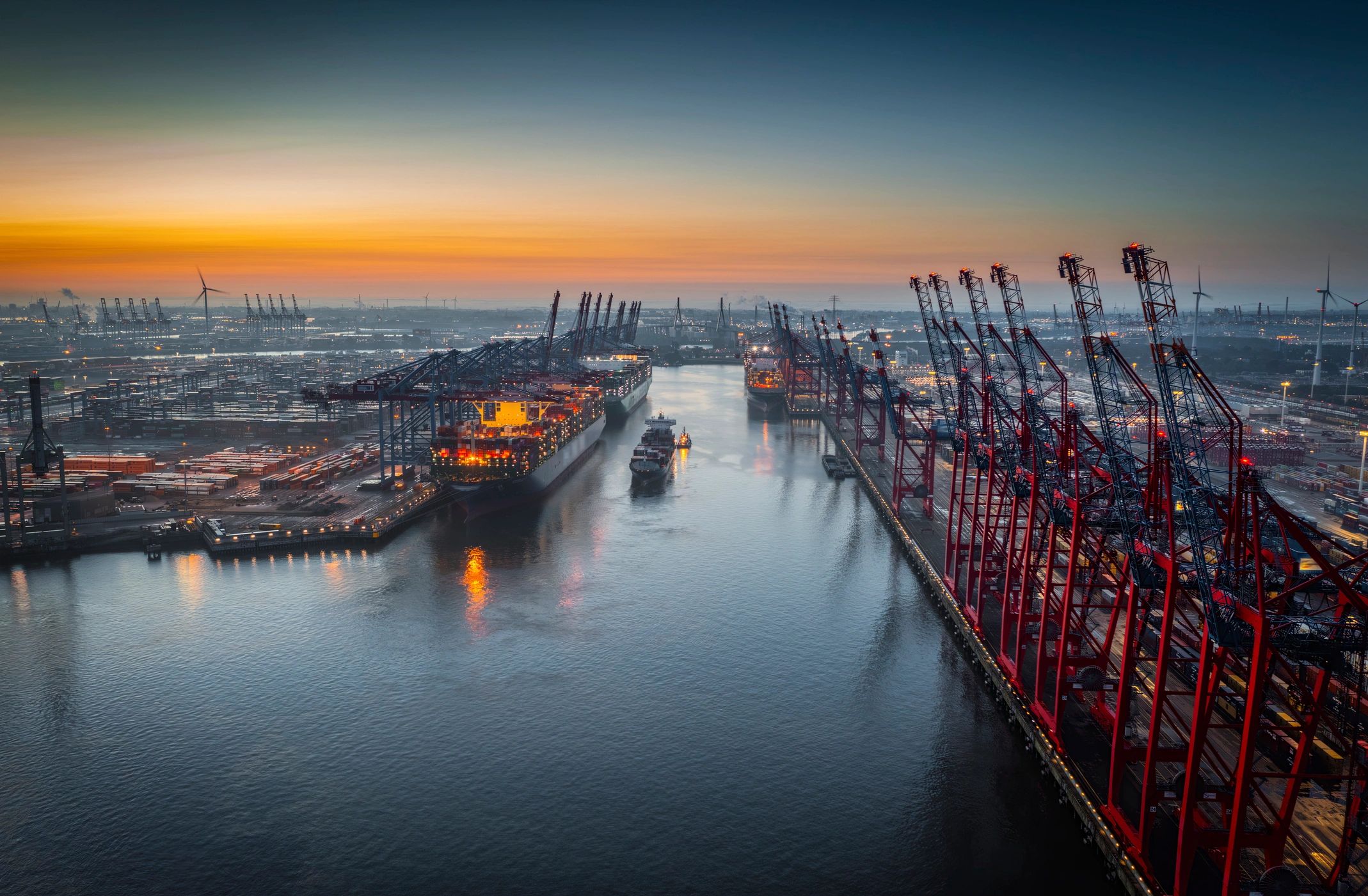 A busy industrial port at sunset with container ships and cranes.