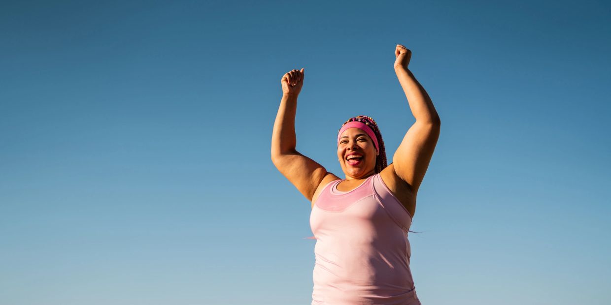 Woman in pink workout outfit raising her arms joyfully outdoors.