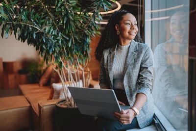 A woman with a laptop sits by a window, looking outside with a smile.