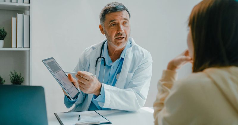 Senior doctor in lab coat using digital tablet discusses medical test results with a patient in clinical office. Medical health care concept.