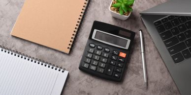 Calculator, notebooks, pen, laptop, and small plant on a desk.