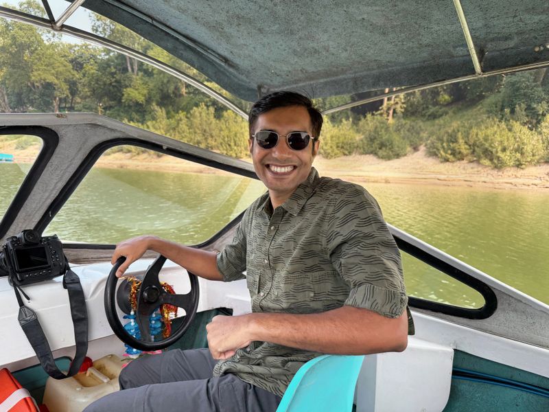 A cheerful male tourist sits on a motorboat gliding through the serene backwaters of Bhitarkanika Mangrove Forest in Odisha, India. Looking directly at the camera with a warm smile, he exudes joy and adventure. The lush greenery of the mangroves and calm waterways form a picturesque backdrop, capturing the essence of eco-tourism and the natural beauty of this biodiverse region. Ideal for illustrating travel, adventure, and the vibrant spirit of exploration in India's unique landscapes.