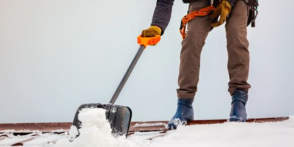 Person shoveling snow off a roof wearing safety gear and gloves.