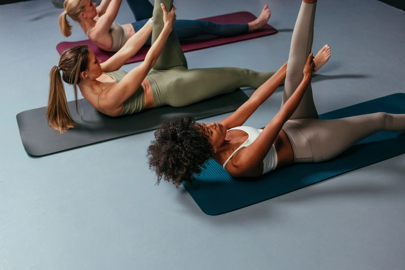 Three young adult women lying on yoga mats, performing stretching exercises for legs and back during pilates class