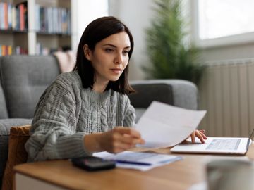 A woman reviewing documents while working on a laptop at home.