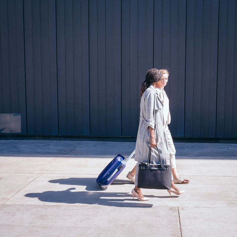 Two women walking side by side pulling luggage on an urban sidewalk with a blue wall background. Ideal for travel, urban lifestyle, or companionship concepts.