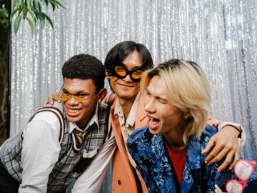 Three friends joyfully posing with quirky glasses in front of a shimmering silver backdrop.