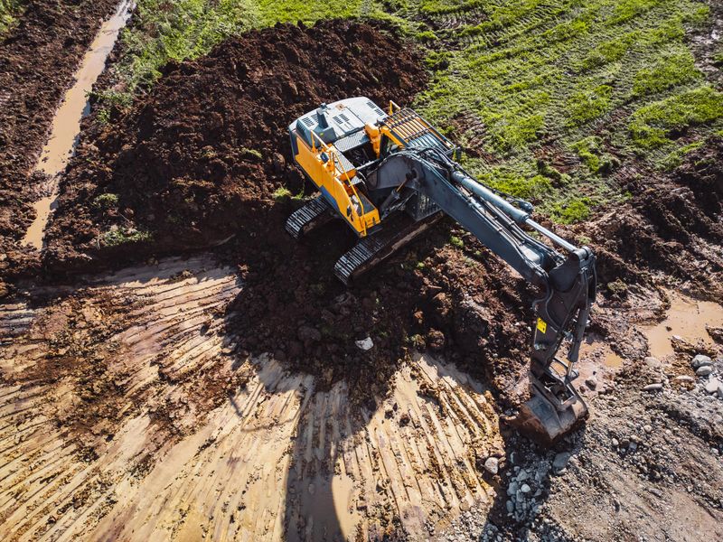 An aerial view of a yellow and black excavator digging into a muddy area, surrounded by freshly turned earth and green grass. The machine is positioned on a construction site, with tracks visible in t